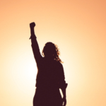 A silhouette with long curly hair raises a fist in the sunlight.