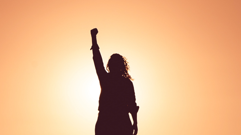 A silhouette with long curly hair raises a fist in the sunlight.