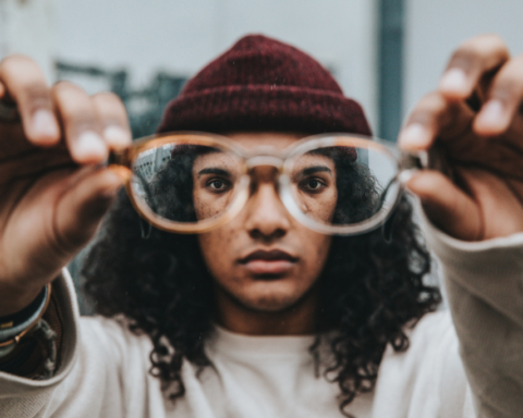 A person of color holds up their eyeglasses to the viewer. You can see their eyes through the frames.