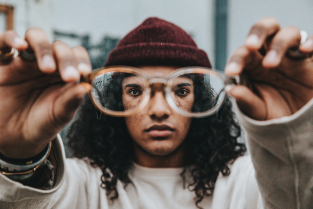 A person of color holds up their eyeglasses to the viewer. You can see their eyes through the frames.