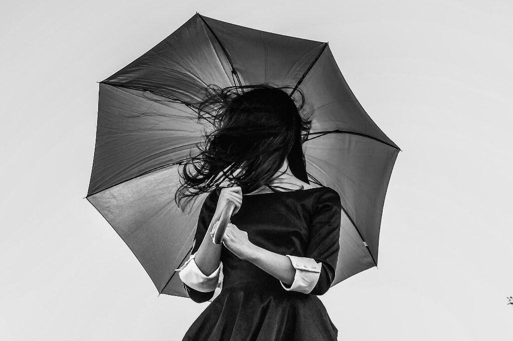 A woman in a dress holds up an umbrella, her hair in her face, in this photograph.