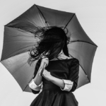 A woman in a dress holds up an umbrella, her hair in her face, in this photograph.