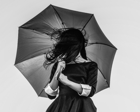 A woman in a dress holds up an umbrella, her hair in her face, in this photograph.