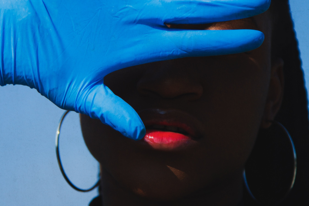 A hand covered in a blue medical glove is raised over a Black person's face.