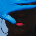 A hand covered in a blue medical glove is raised over a Black person's face.