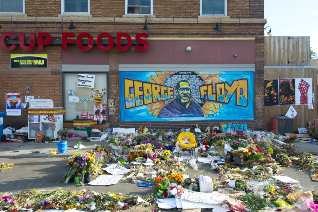 The George Floyd memorial at his place of death is strewn with flowers, notes, and a mural of his face and name.