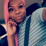 A photograph of Muhlaysia Booker, a Black woman with pink hair and a blue and white shirt in her car.