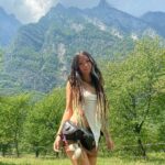 An image of Shani Louk, a 23-year-old woman with long dread locks and wearing a white tank top and black hip-bag, standing in front of a mountain range.