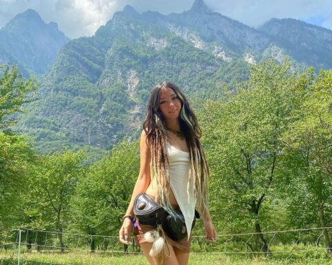 An image of Shani Louk, a 23-year-old woman with long dread locks and wearing a white tank top and black hip-bag, standing in front of a mountain range.