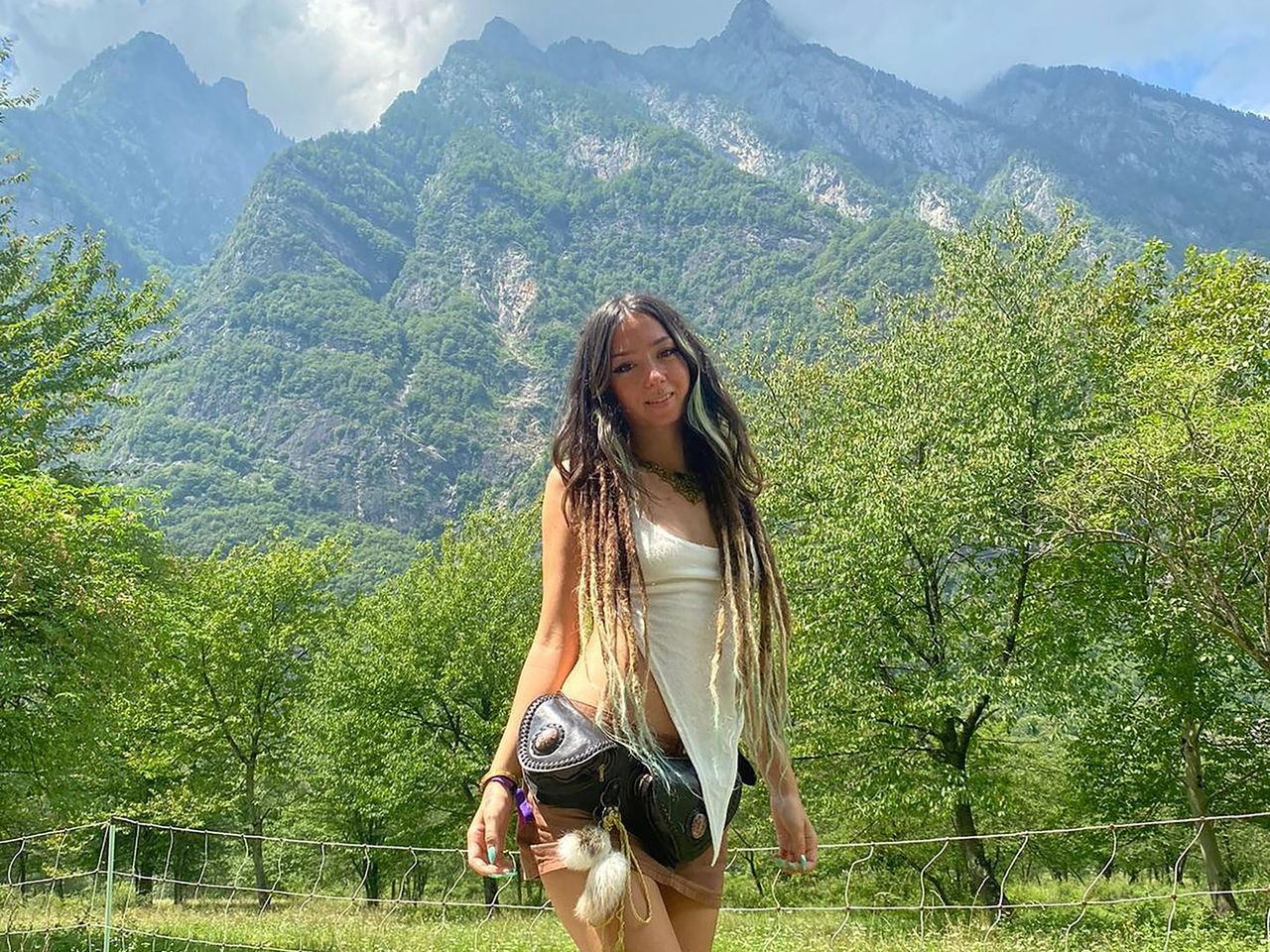 An image of Shani Louk, a 23-year-old woman with long dread locks and wearing a white tank top and black hip-bag, standing in front of a mountain range.