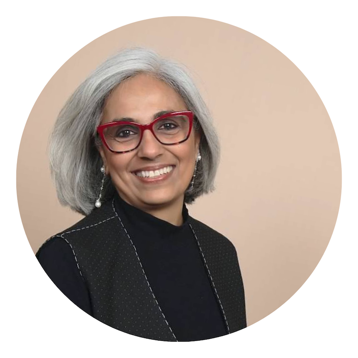 Photo of Kashiana Singh, a woman with silver hair, red glasses and a black coat, against a beige background.