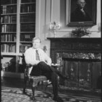 A photograph of Jimmy Carter in the white house, seated on a chair in front of a portrait of George Washington.