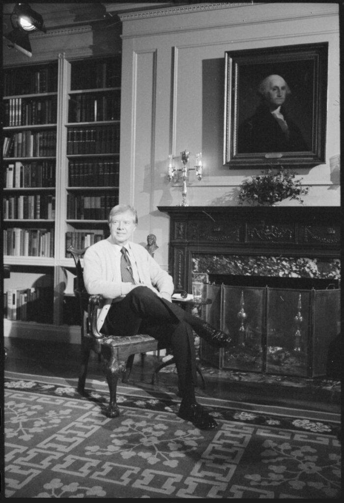 A photograph of Jimmy Carter in the white house, seated on a chair in front of a portrait of George Washington.