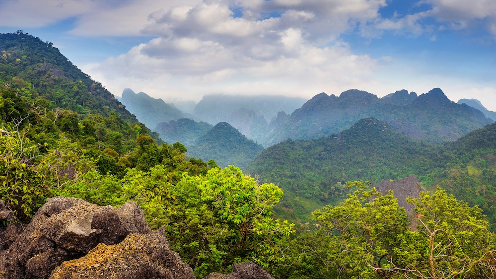 A Laotian mountain-side covered in verdure below a sky full of light clouds.